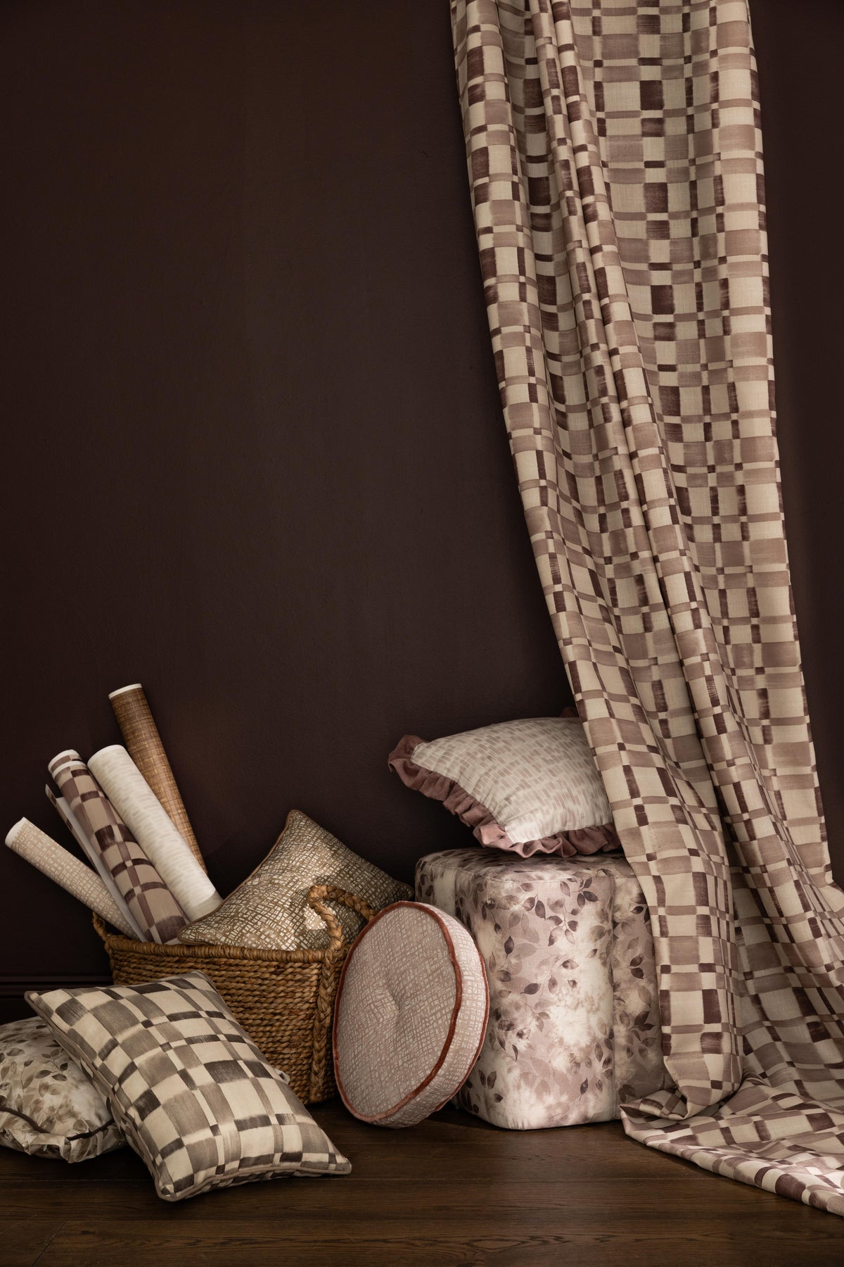 Styled interior scene with draped fabric with a brown checkerboard along side stacked throw pillows, ottoman and basket with wallpaper rolls in various patterns in a range of muted colors against a dark brown wall. 