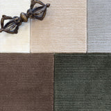 Close up of broadloom carpet samples with solid cut pile ribbed stripe in white, beige, grey brown and green with an ornamental brass sculpture. 
