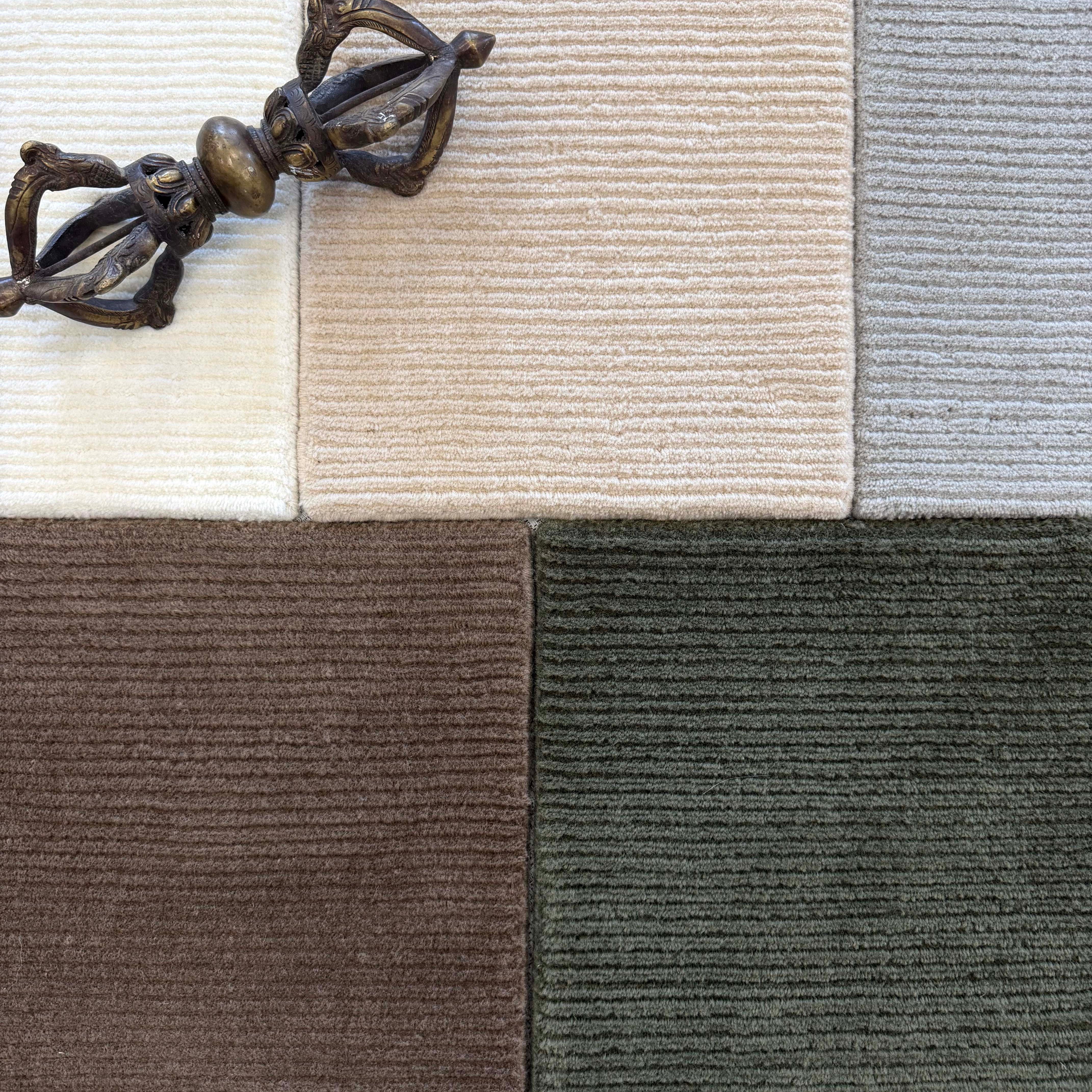 Close up of broadloom carpet samples with solid cut pile ribbed stripe in white, beige, grey brown and green with an ornamental brass sculpture. 