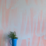 A vase of flowers stands in front of a wall papered in an abstract paint print in light blue on a pink field.