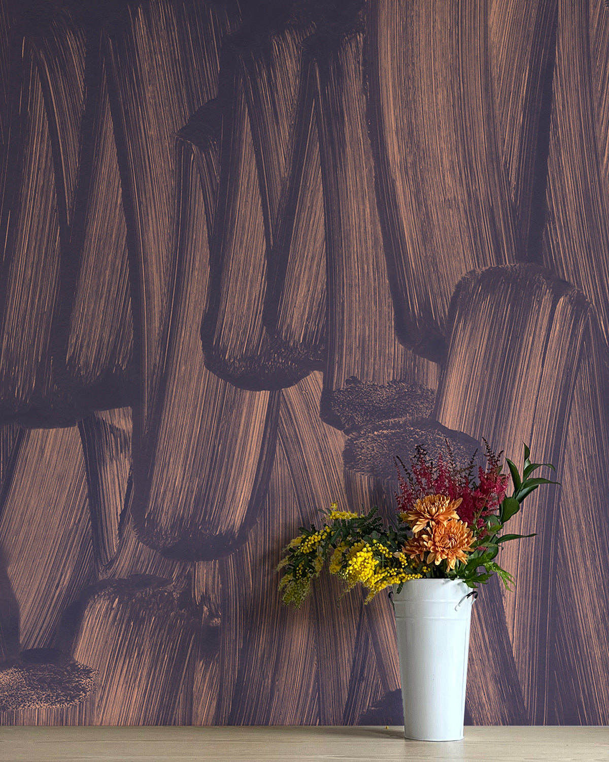 A vase of flowers stands in front of a wall papered in an abstract paint print in dark grey on a purple field.