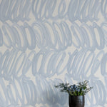 A vase of flowers stands in front of a wall papered in a playful paint print in blue on a light blue field.