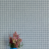 A vase of flowers stands in front of a wall papered in a dense plaid pattern in shades of blue-gray.