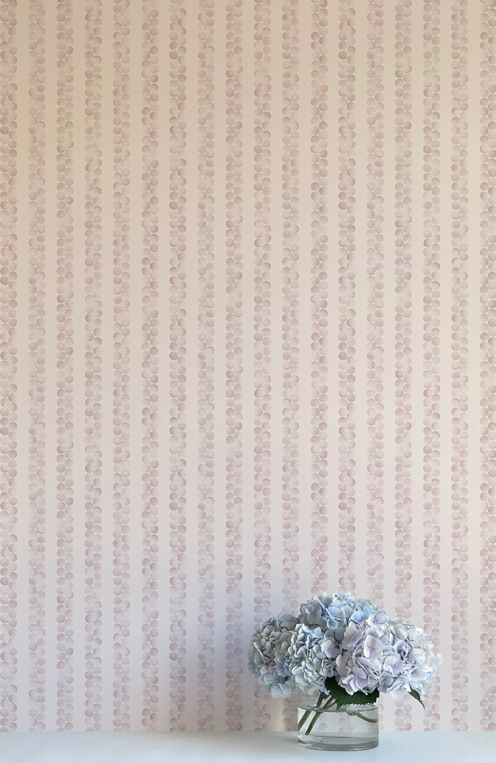 A vase of flowers stands in front of a wall covered in a repeating striped circular print in purple on a cream field.