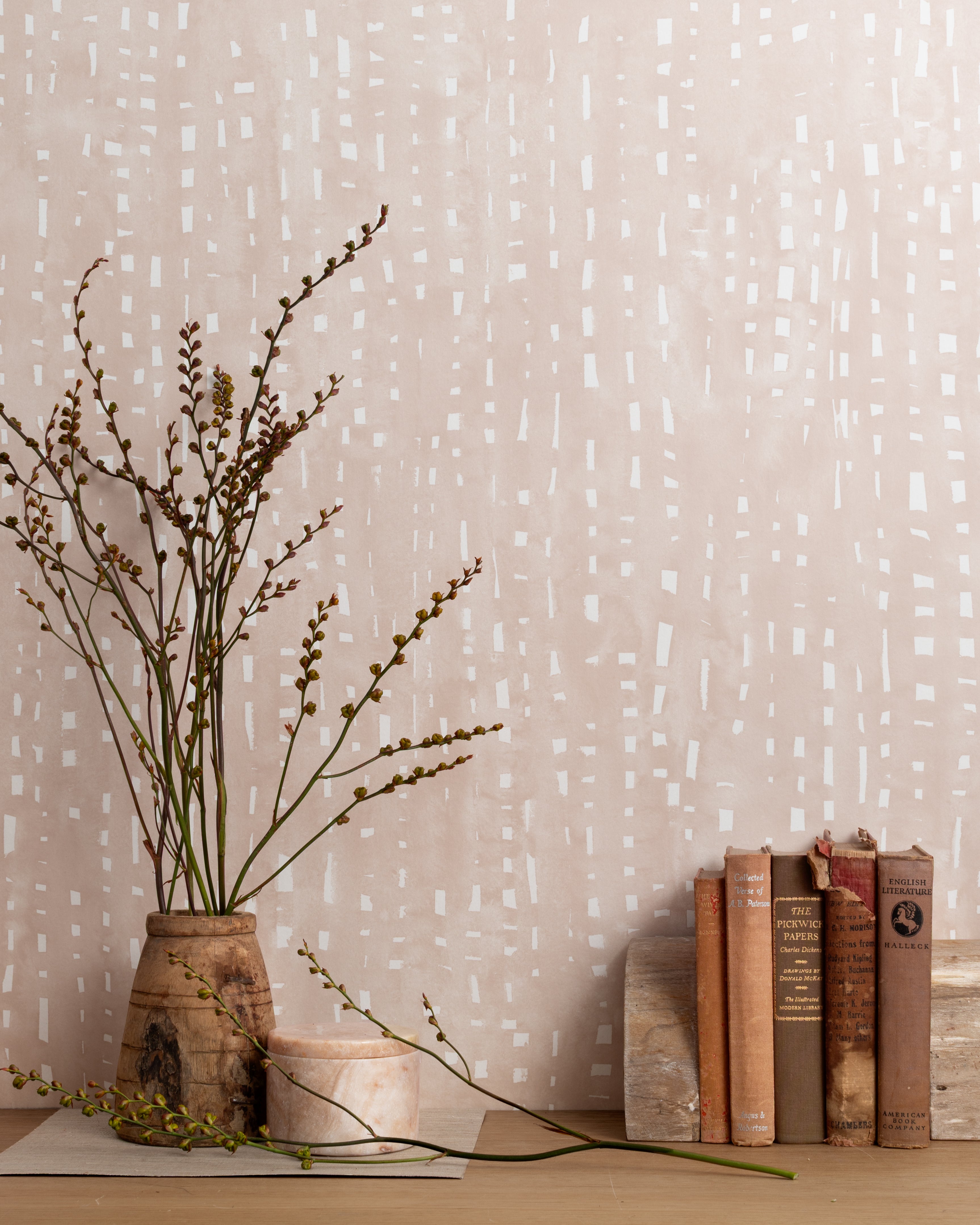 Mantle vignette with vase and brances and antique books in front of a wall papered in a painterly dashed dot print in white on a pale pink field.