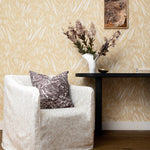 Cream slip covered chair with grey wave print throw pillow against a console table with flowers against wallpaper in abstract leaf pattern in white on a pastel yellow watercolor field.