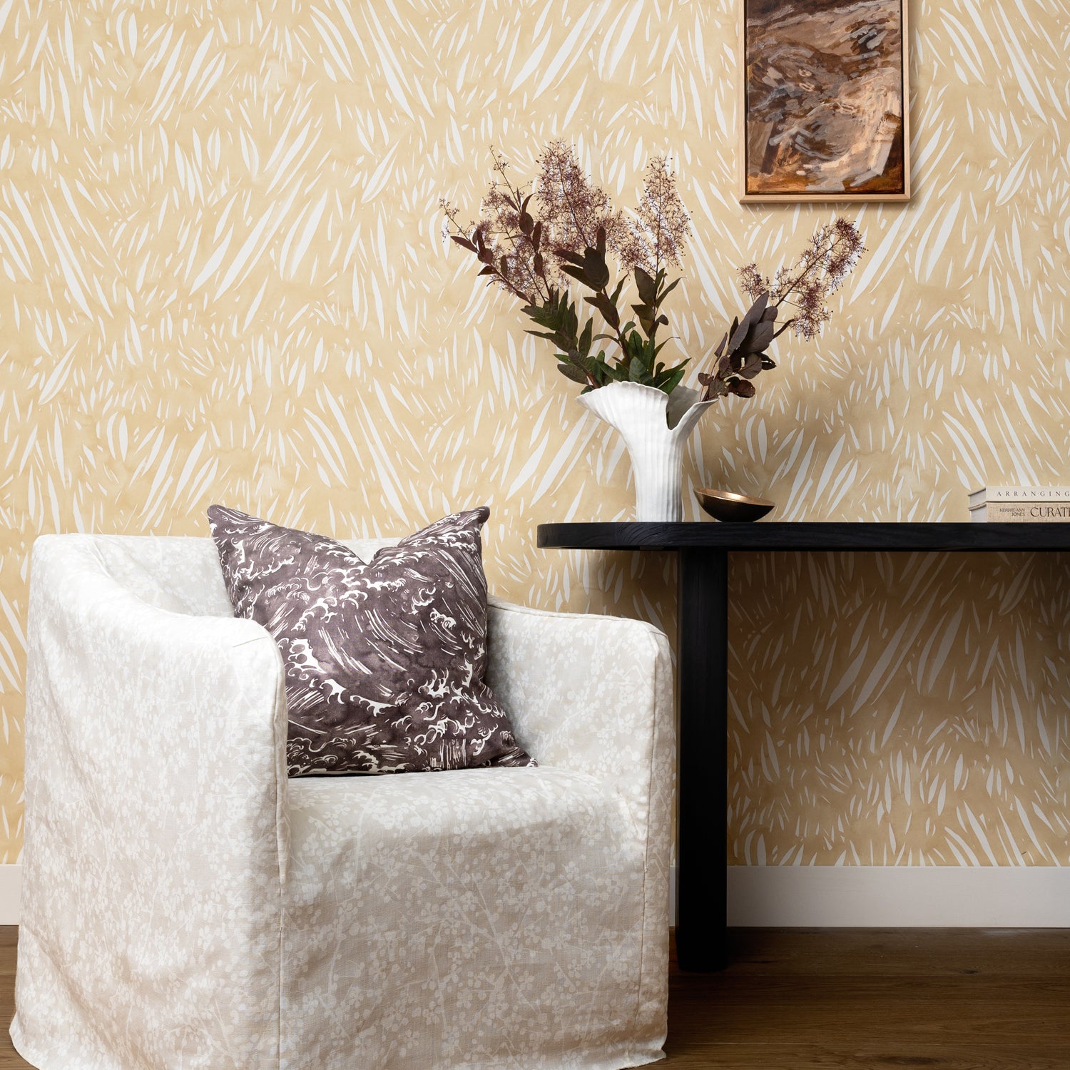 Cream slip covered chair with grey wave print throw pillow against a console table with flowers against wallpaper in abstract leaf pattern in white on a pastel yellow watercolor field.
