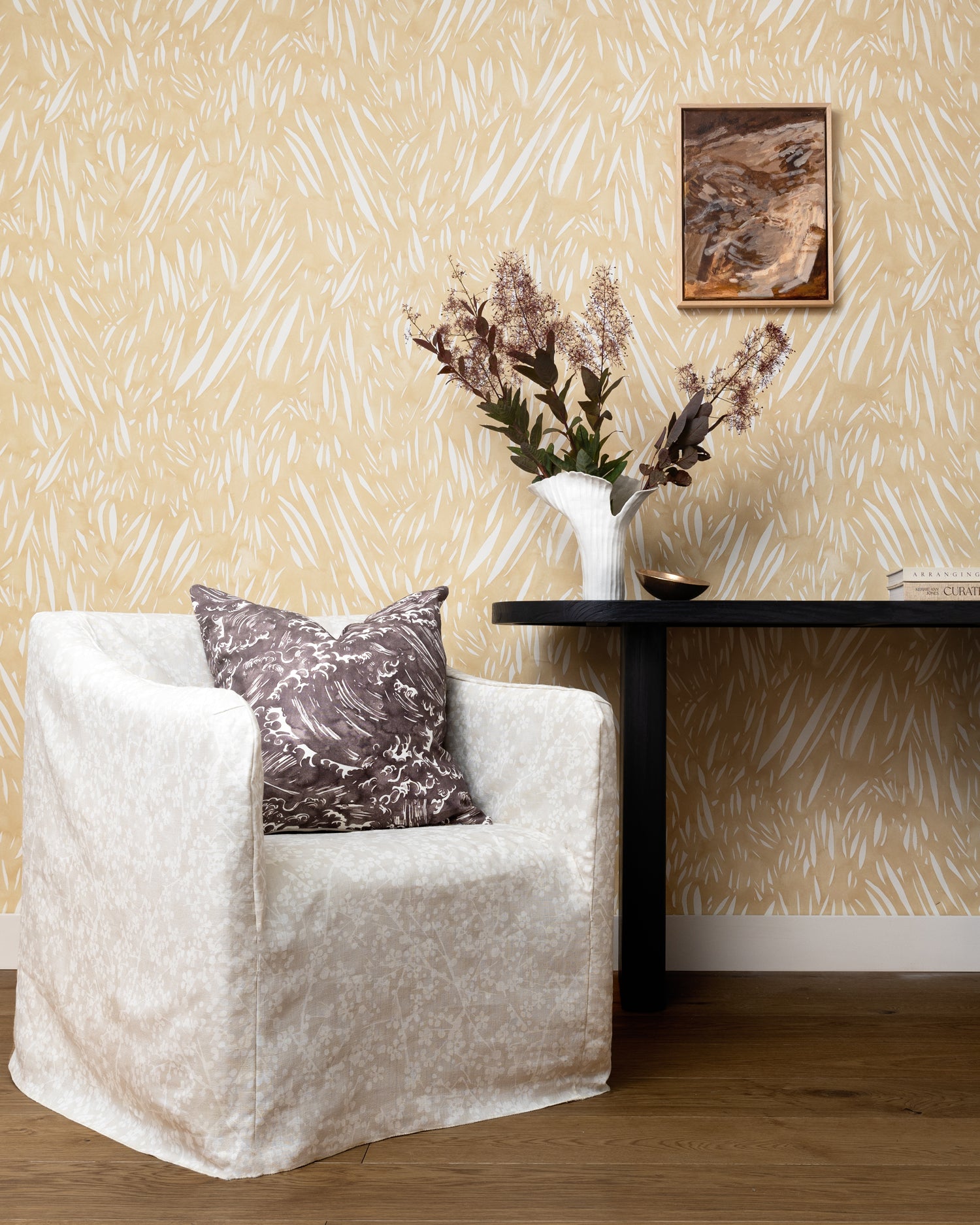Cream slip covered chair with grey wave print throw pillow against a console table with flowers against wallpaper in abstract leaf pattern in white on a pastel yellow watercolor field.