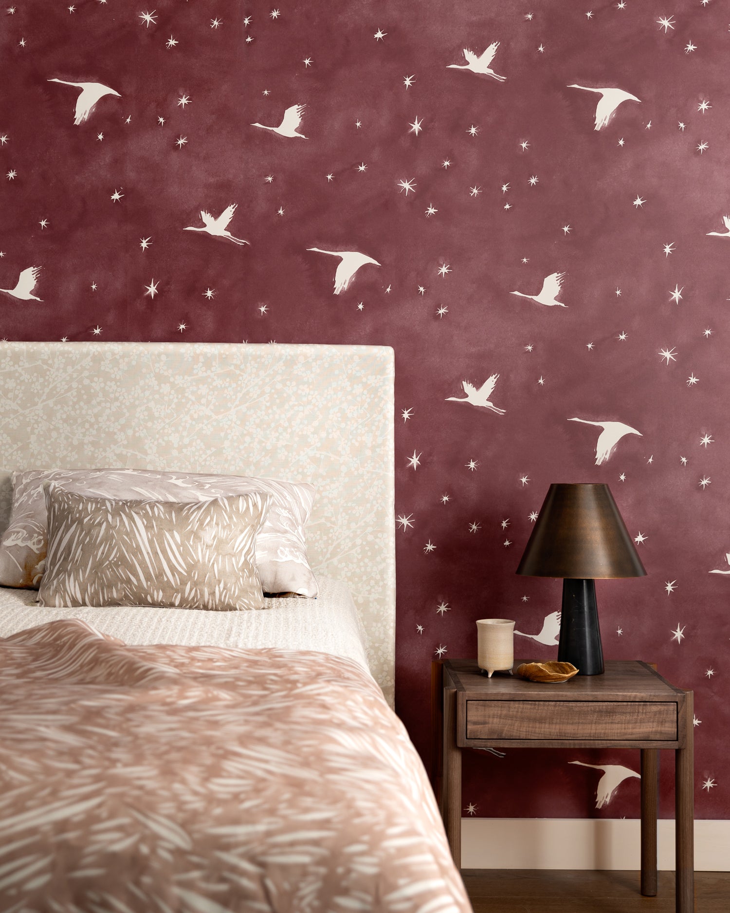 Bedroom scene with layered printed bed clothes in shades of taupe and pink against a wallpaper with stars and flying geese in white on a crimson watercolor field.