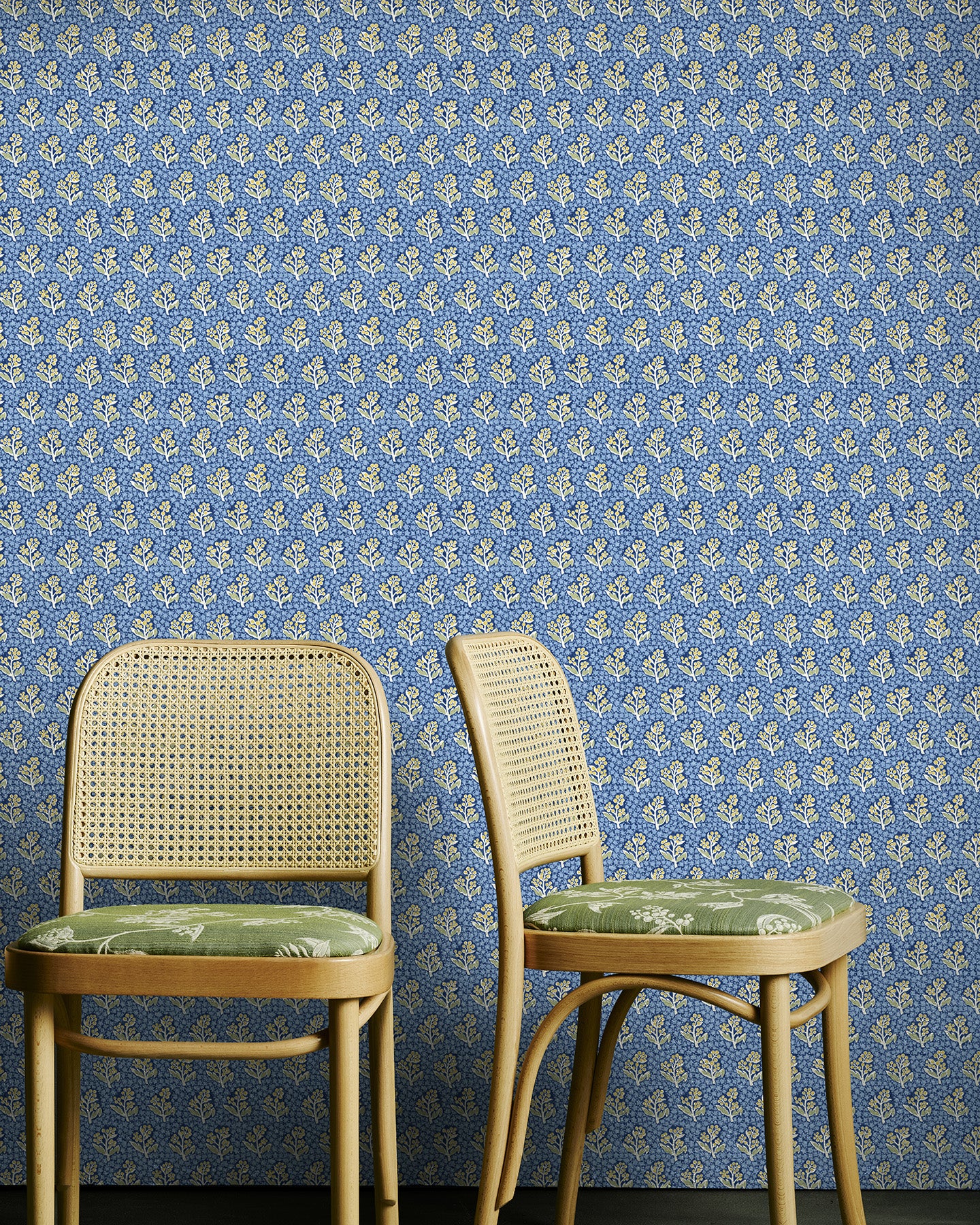 Two wicker chairs with green cushions against a blue floral wallpapered wall.