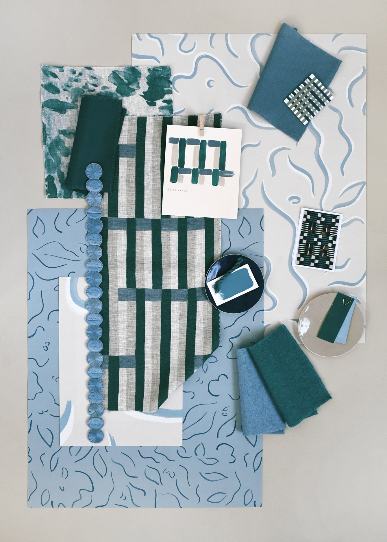 Layered mood board with printed fabrics, wallpapers, trims and swatches in shades of teal, blue, taupe and cream.