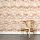 A wooden chair stands in front of a wall papered in a painterly ikat print in shades of tan and orange on a white field.
