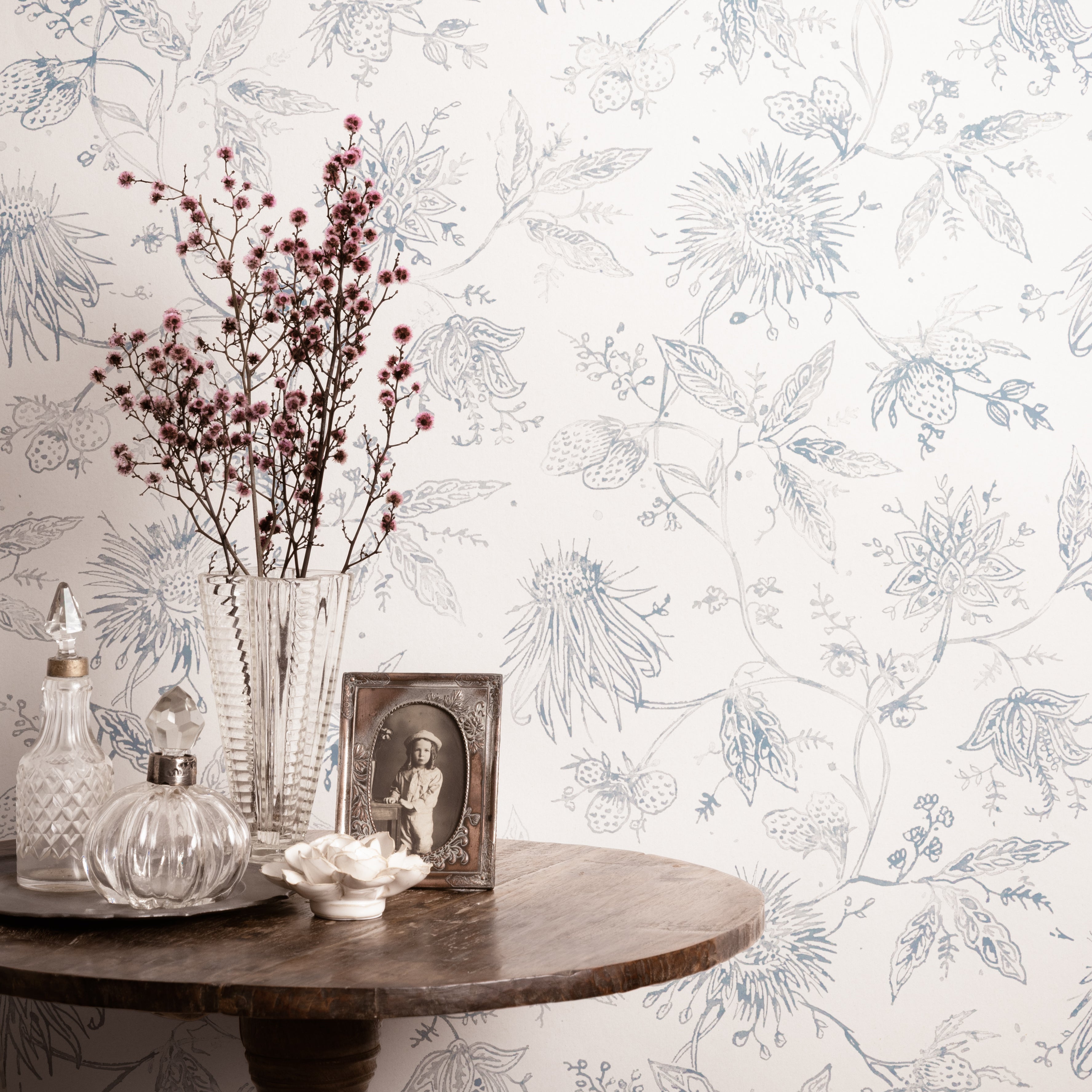 Decorative table with a vase, bottles, and photo frame against a floral wallpapered wall.