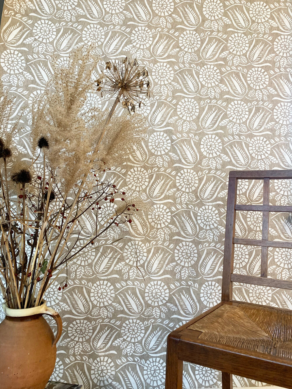 A chair and bunch of flowers stands in front of a wall papered in a dense floral print in white on a tan field.