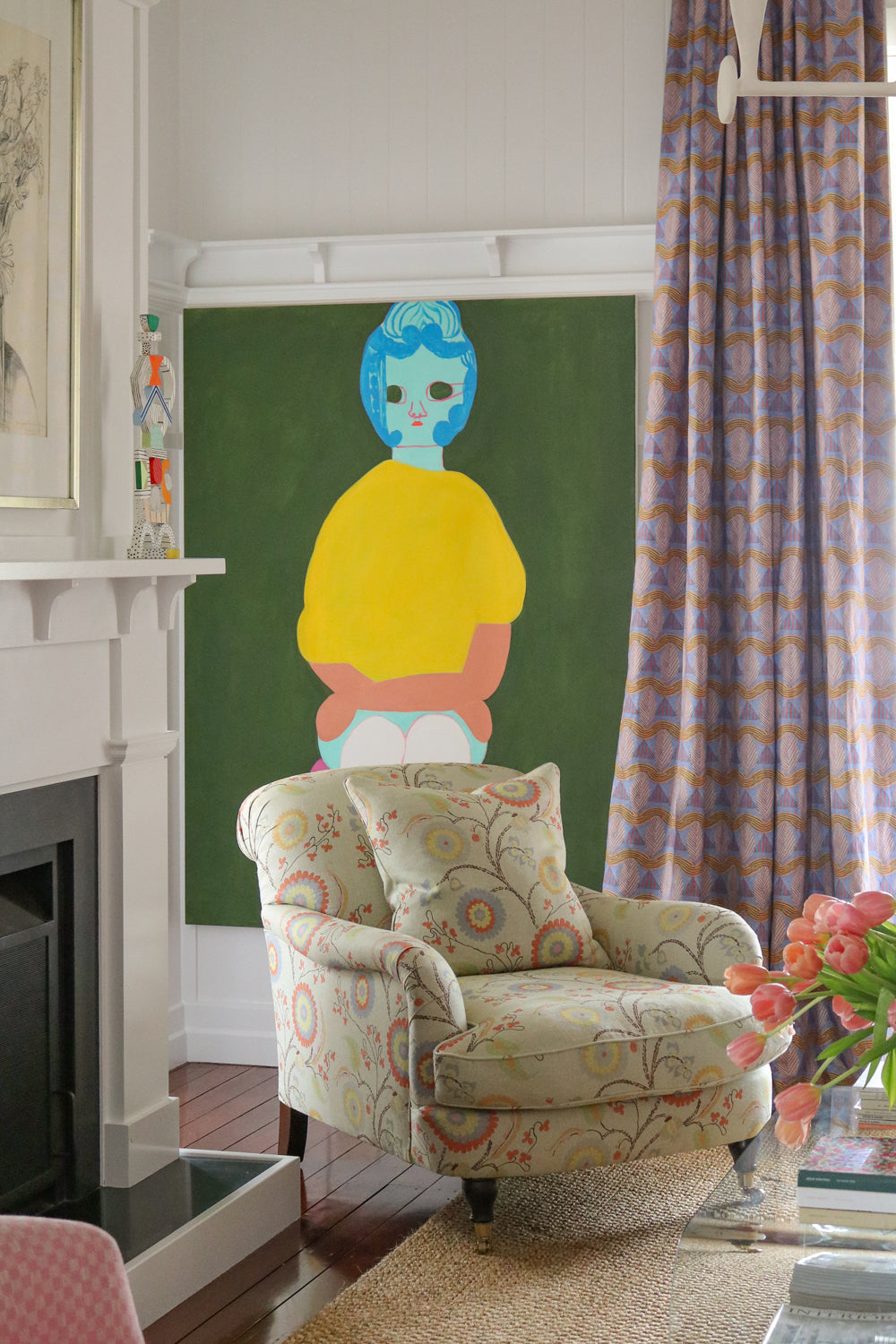 Living room interior with white fire place mantle.  pastel floral upholstered arm chair against a painting of a woman's abstracted figure on a green field with a purple geometric curtain.