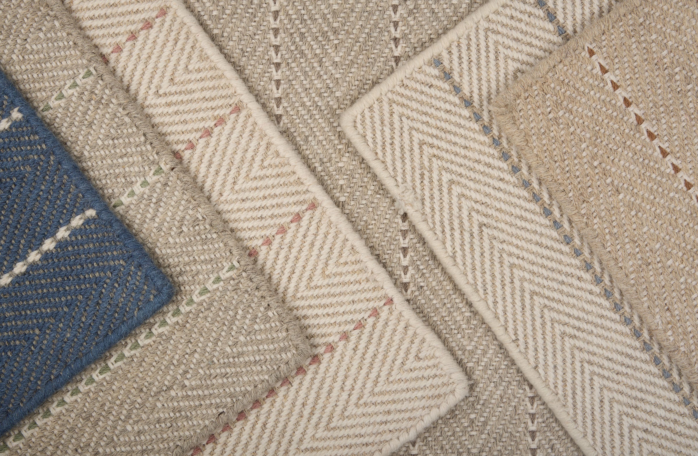 Stack of broadloom carpet samples in a striped herringbone design in an array of netural colors with accents of blue, pink, brown, green and grey. 