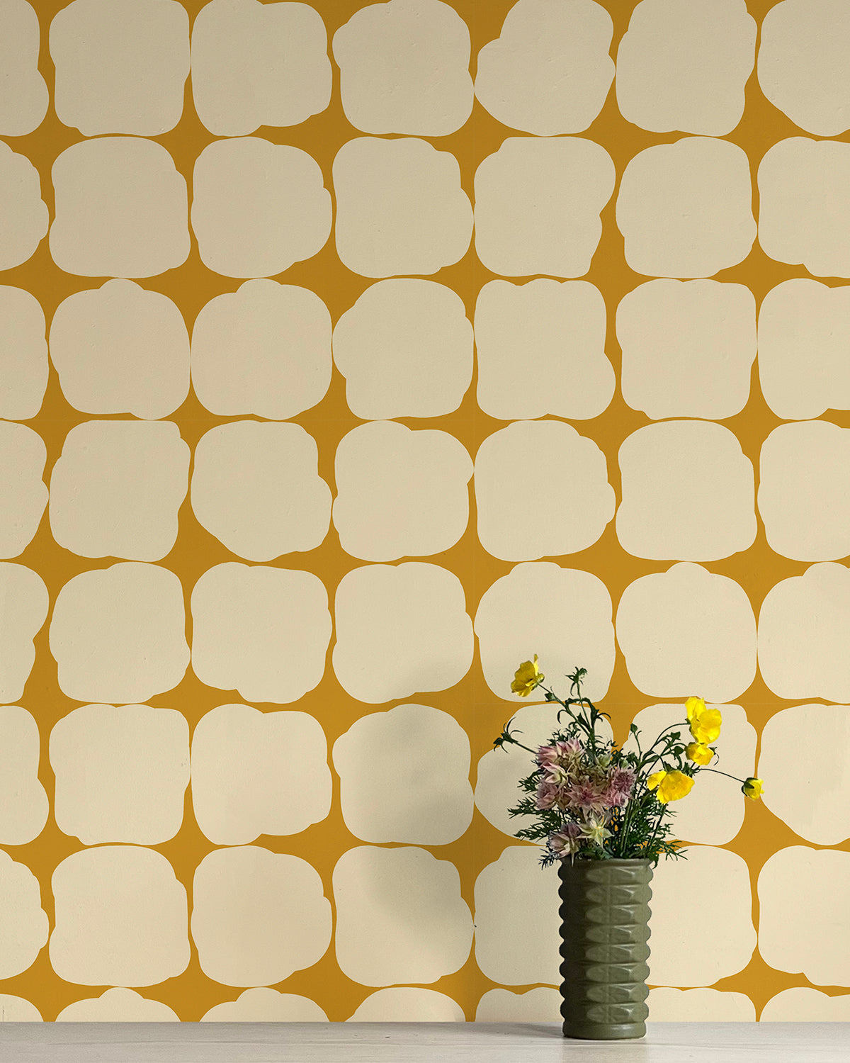 A vase of flowers stands in front of a wall papered in a repeating blot print in cream on a yellow field.