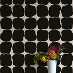 A vase of flowers stands in front of a wall papered in a repeating blot print in black on a white field.