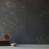 A table with knick knacks stands in front of a wall papered in a random splattered pattern in metallic gold on a black field.