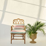 A rattan armchair with a striped white, pink and green cushion sits next to a potted palm plant against a white wall with window shadow patterns.