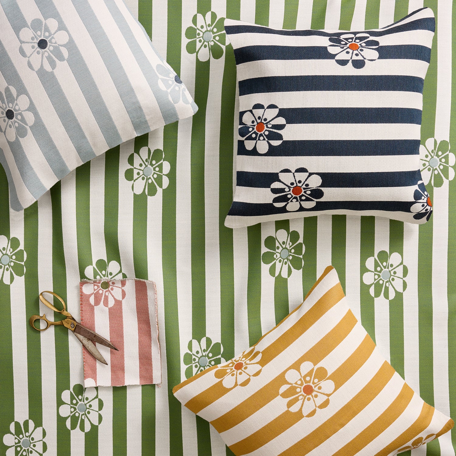 Three decorative throw pillows with striped and floral patterns are arranged on a green and white striped fabric background, alongside a small pair of scissors and a folded matching fabric swatch.