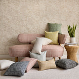 A styled interior scene with a blush pink upholstered settee surrounded by multiple decorative throw pillows in neutral and muted tones. A small wicker side table with a potted snake plant sits to the right against a textured beige wallpaper background.
