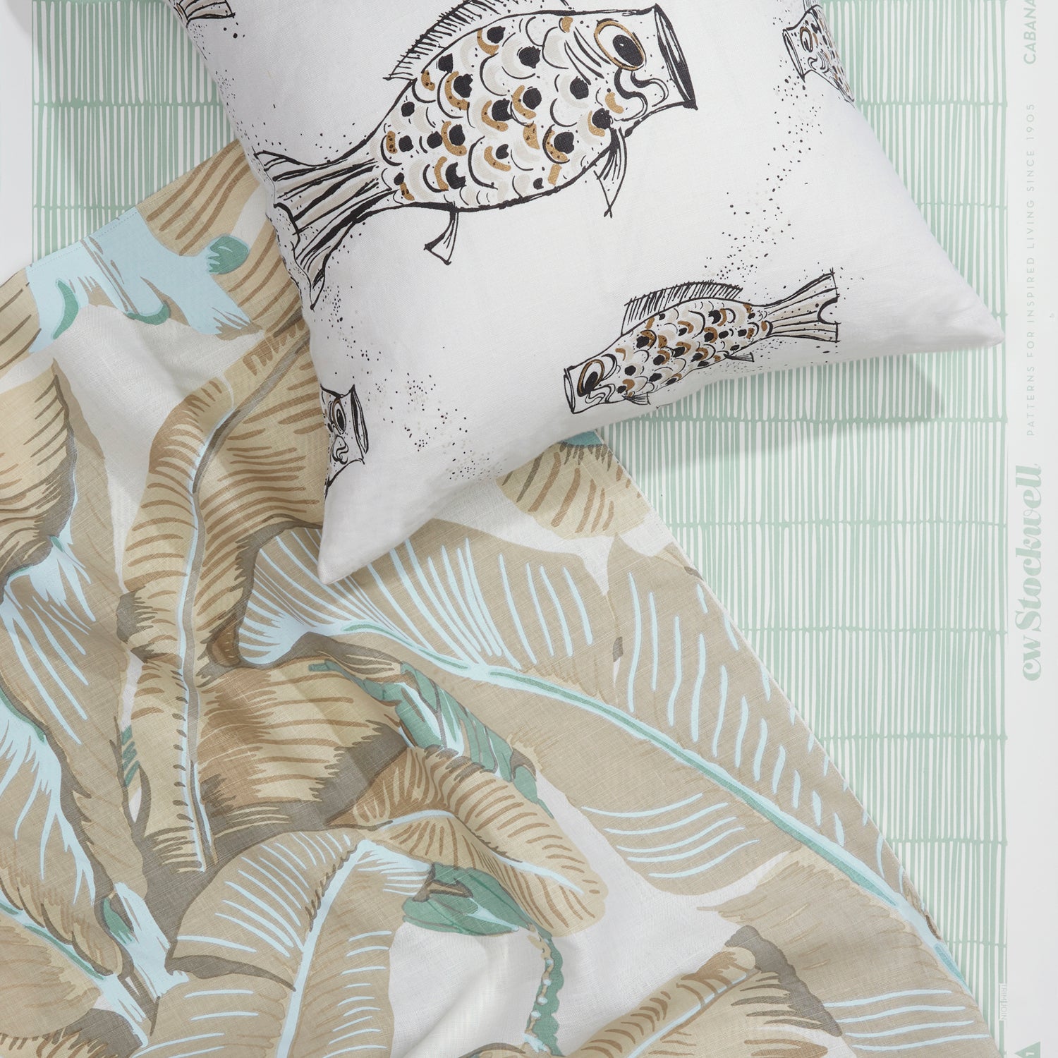 A styled flat-lay with banana leaf pattered fabric in tan and seafoam, with throw pillow with black and white fish pattern atop a wallpaper with a bamboo stripe pattern in aqua blue and white. 