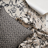 A styled flat-lay image with a throw pillow in a small scale black and white lattice design layered over a hand drawn branch and flower fabric and wallpaper in black, white and beige. 