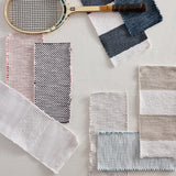 A styled flat-lay of multiple woven fabric swatches in neutral and muted tones, including a tennis racket and folded textile pieces, arranged on a light textured background.