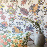 A close-up of a floral wallpaper with a vintage botanical pattern in orange, yellow, blue, and green on a white background, styled with a matching floral throw pillow and a textured gray vase.
