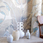 A styled interior scene showing a set of white ceramic vases and a textured beige vase arranged on a stone console table in front of a tropical leaf patterned wallpaper and curtain.