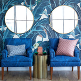 Two blue upholstered armchairs with patterned pillows are placed in front of a tropical leaf wallpaper wall with two round gold-framed mirrors. A small gold side table with green plants and a pink box sits between the chairs.