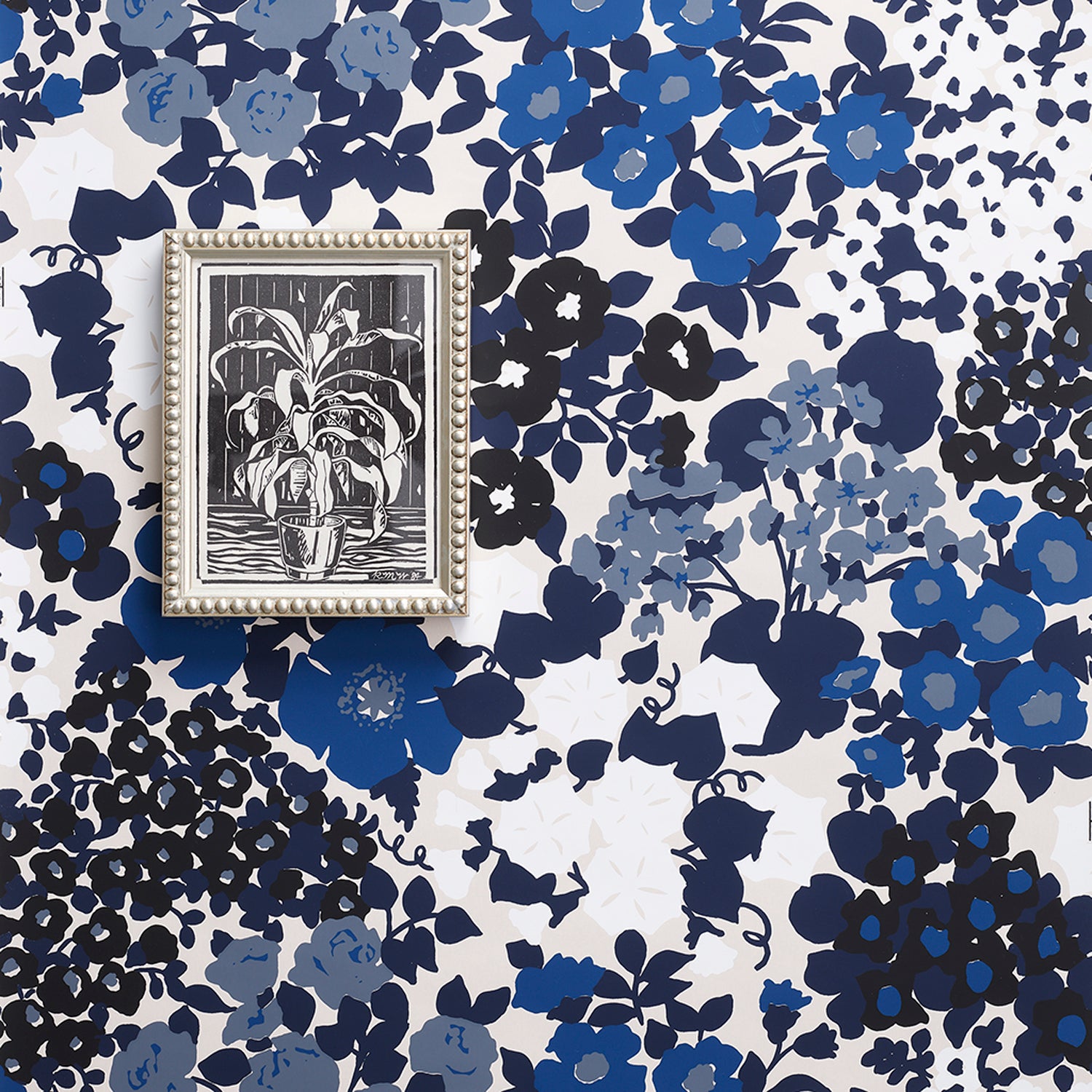 A small framed black-and-white botanical artwork with a silver-toned ornate frame, placed on top of a blue, navy, and white floral wallpaper pattern.