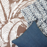 A styled flat-lay of home textiles featuring a brown and white palm leaf patterned wallpaper a blue and white leopard print fabric, and a blue bamboo stripe pillow.