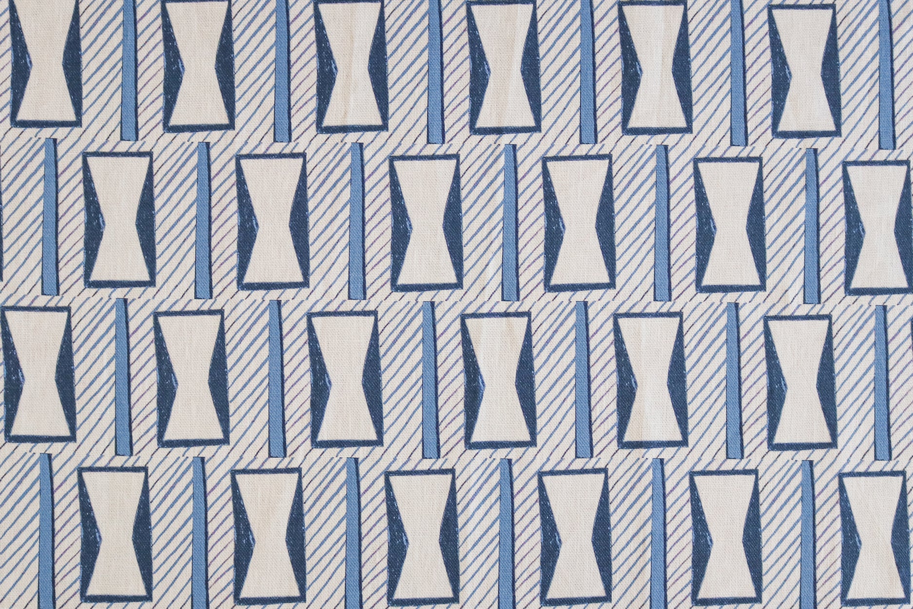 A close-up of a patterned fabric featuring a repeating geometric design of white hourglass-like shapes outlined in blue, set against a light gray background with diagonal blue stripes.