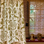 A curtain in a printed fabric in a lion, mixed animal and plant pattern in olive on a white field, against a window with with a reed shade and ceramic vases on the sill. 