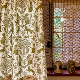 A curtain in a printed fabric in a lion, mixed animal and plant pattern in olive on a white field, against a window with with a reed shade and ceramic vases on the sill. 