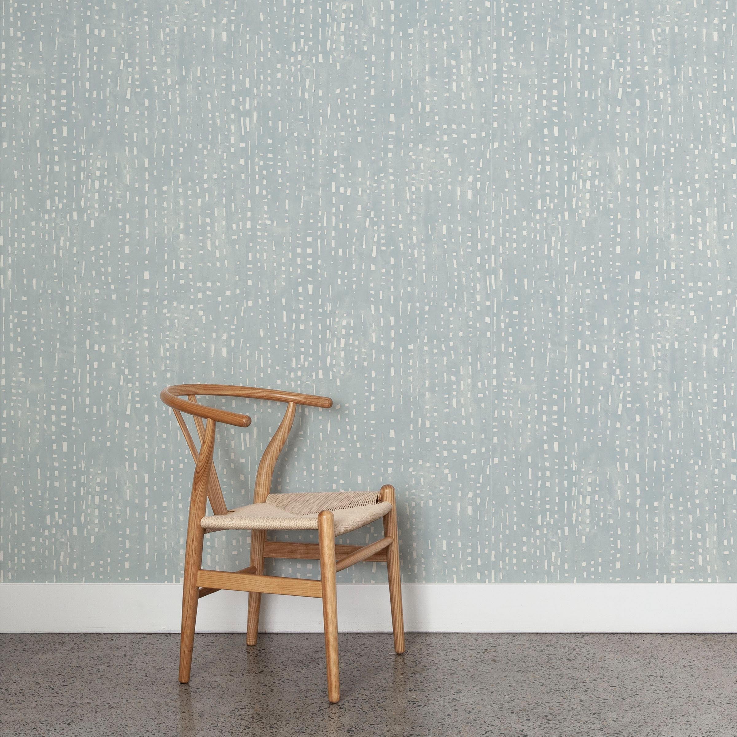 A wooden chair stands in front of a wall papered in a painterly dashed dot print in white on a blue-gray field.