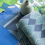 A close-up styled flat-lay of patterned fabric pieces in blue, navy, and teal tones, with a small pale blue ceramic vase placed on top over a colorful floral paper backdrop.
