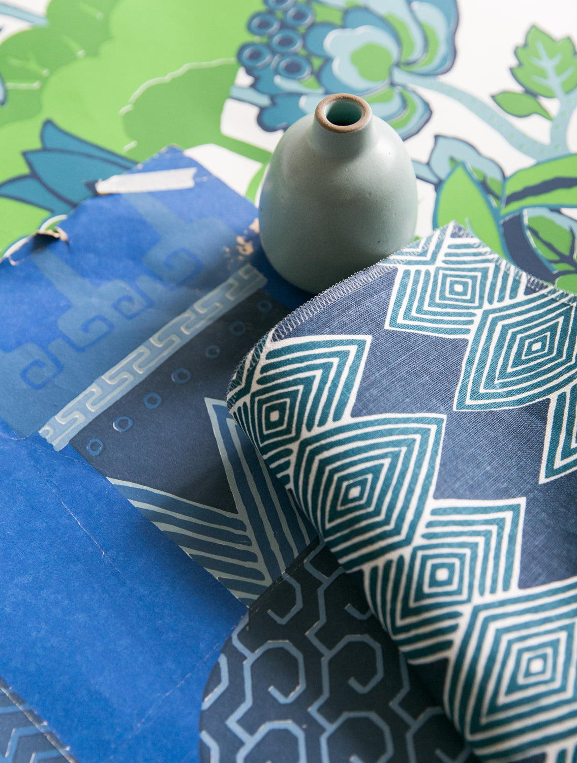 A close-up styled flat-lay of patterned fabric pieces in blue, navy, and teal tones, with a small pale blue ceramic vase placed on top over a colorful floral paper backdrop.