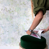 A woman opens a catalog in front of a wall papered in a watercolor painted print in shades of green, yellow and white.