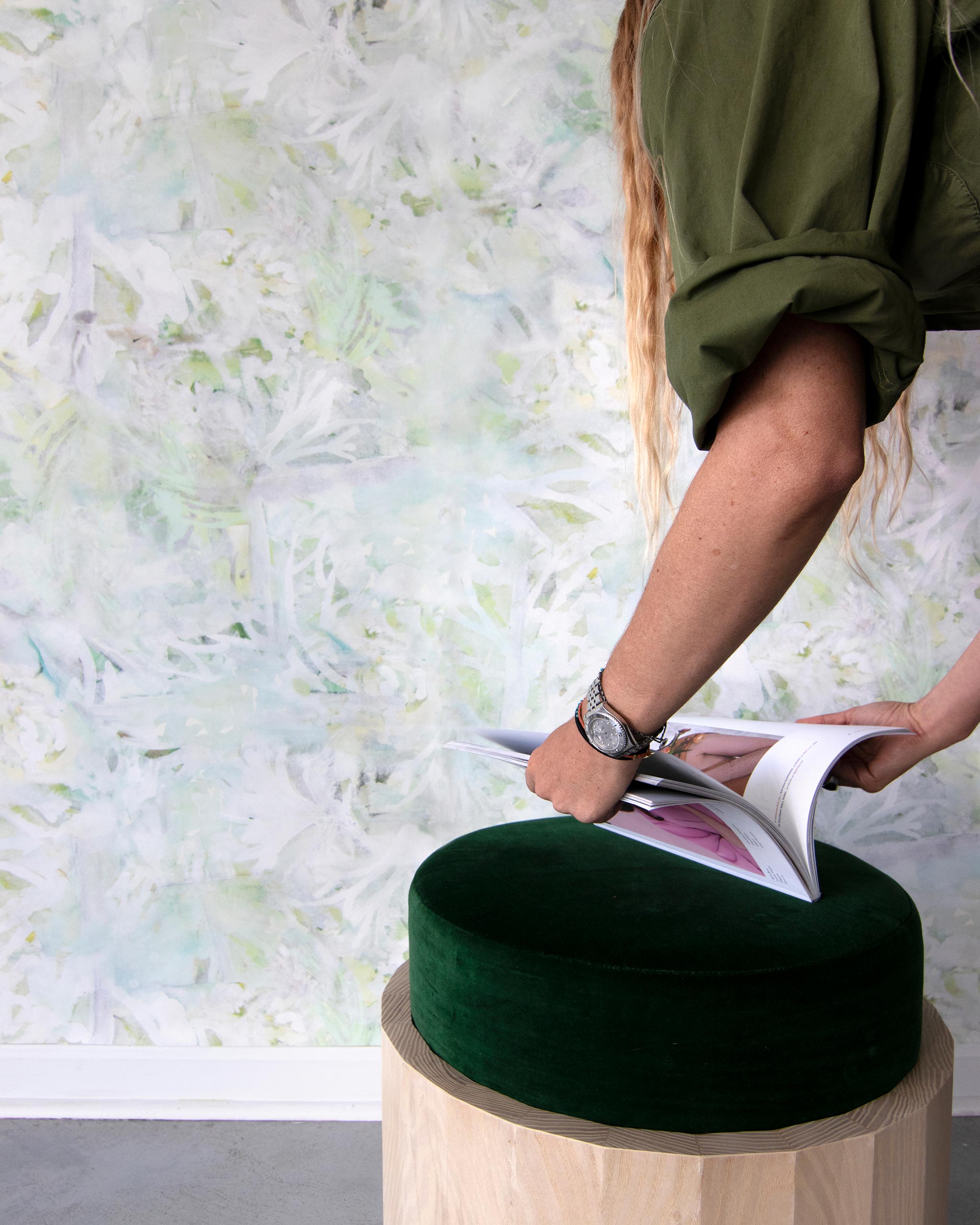 A woman opens a catalog in front of a wall papered in a watercolor painted print in shades of green, yellow and white.
