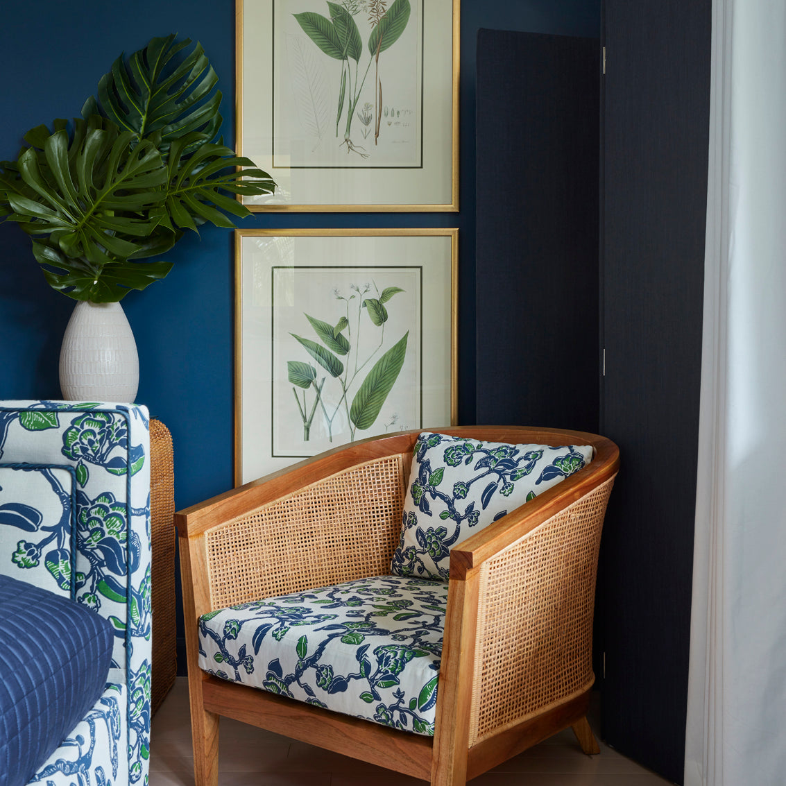 A wooden armchair with blue and white floral patterned cushions, set in a room with a dark blue wall, framed botanical prints, a potted monstera plant, and a black folding screen.