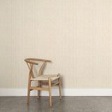 A wooden chair stands in front of a wall papered in an intricate striped grid pattern in gold on a white field.