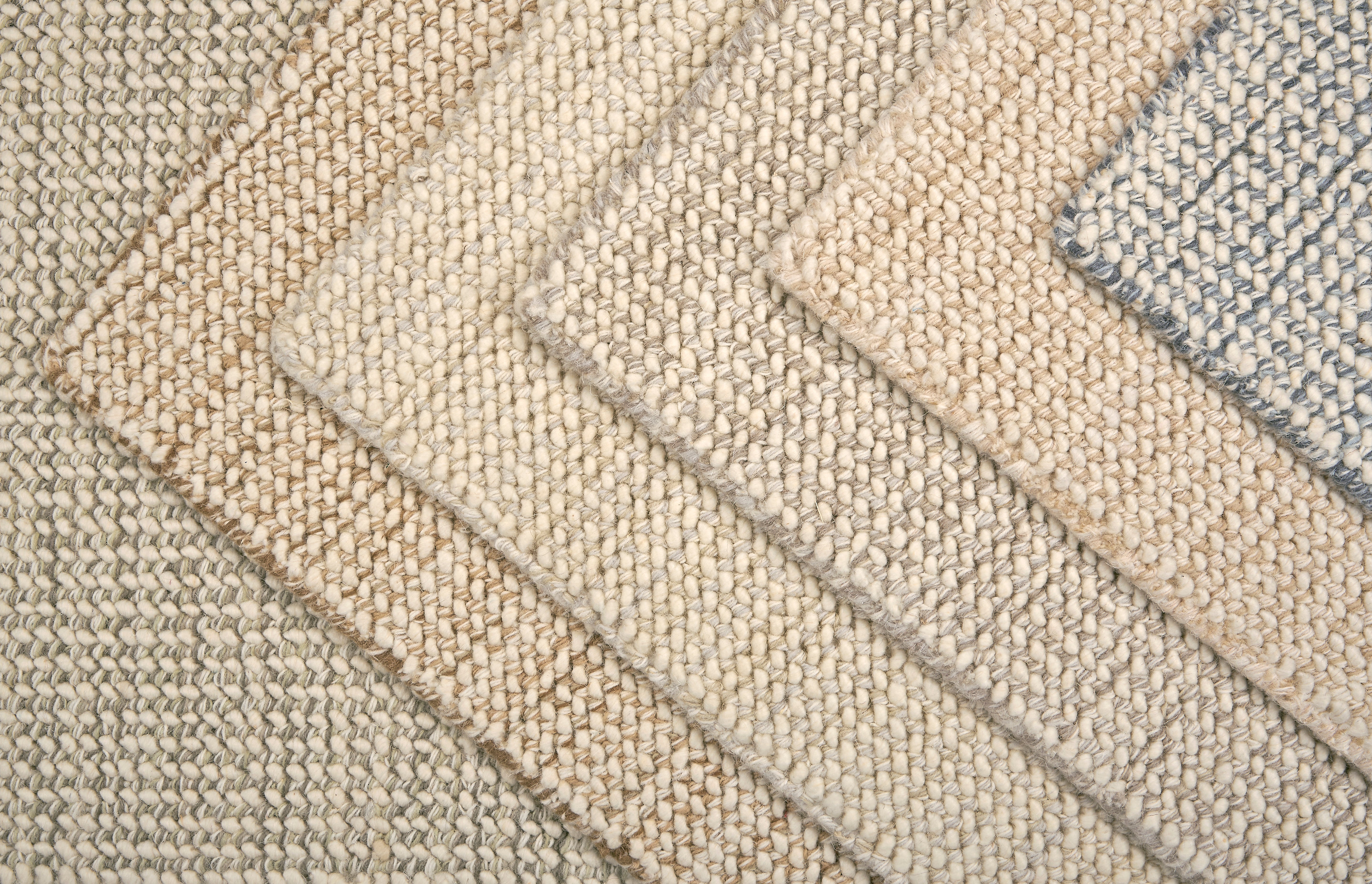 A close-up of overlapping woven rug samples in neutral tones, including beige, cream, tan, and a muted blue-gray, showing the texture and pile of the rugs.