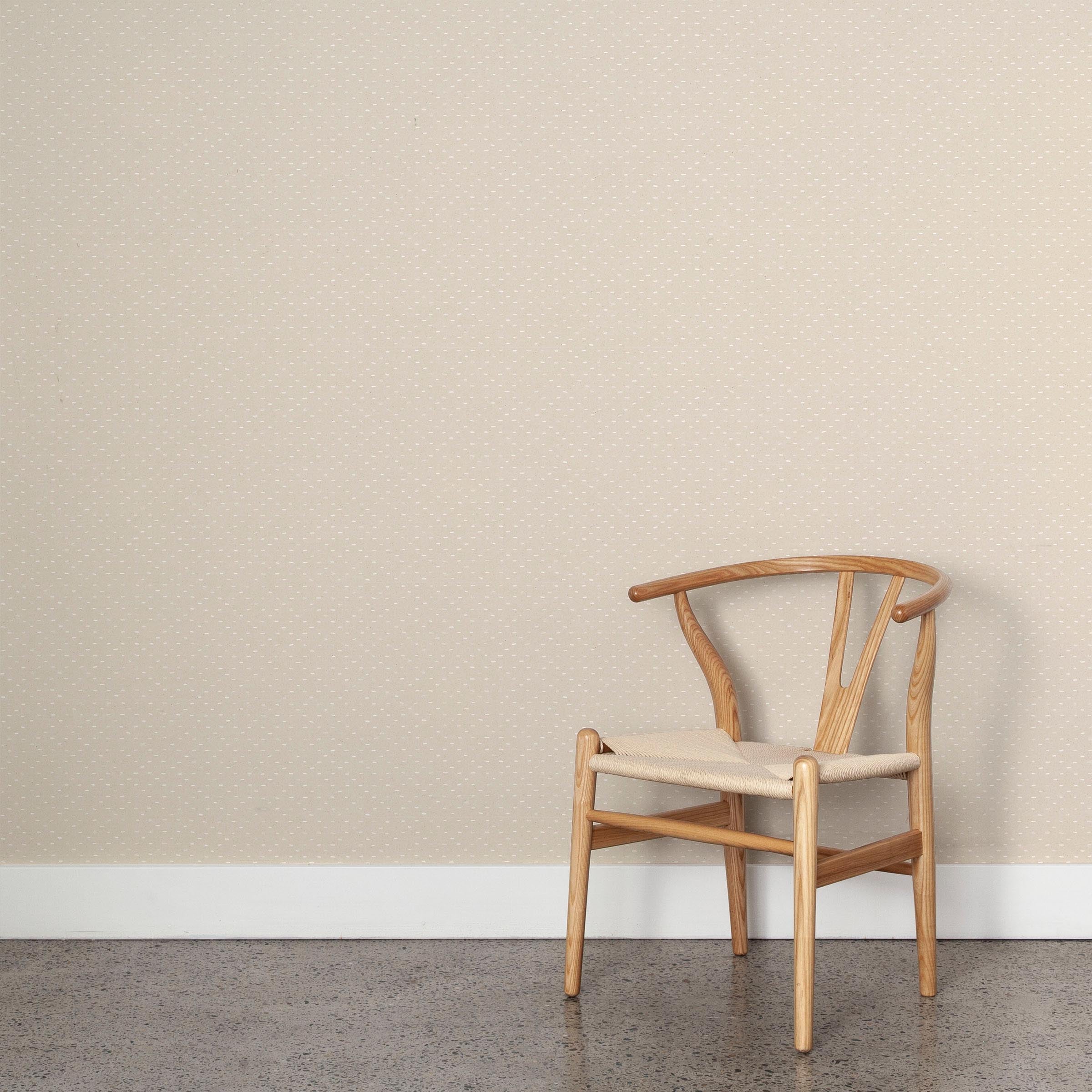 A wooden chair stands in front of a wall papered in a dotted diamond grid in white on a cream field.