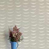 A vase of flowers stands in front of a wall papered in an abstract crescent print in cream on a gray field.