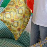 Woman in white tee shirt and jeans standing holding printed geometric pillows against a green checkerboard patterned arm chair.