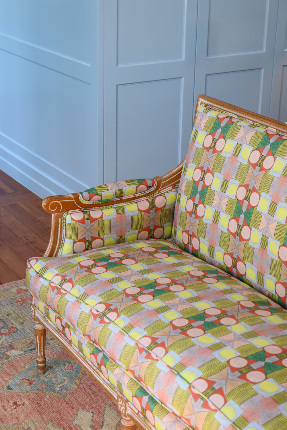 Detail of a wooden settee with bold geometric patterned upholstery on a Persian rug against a blue paneled wall . 