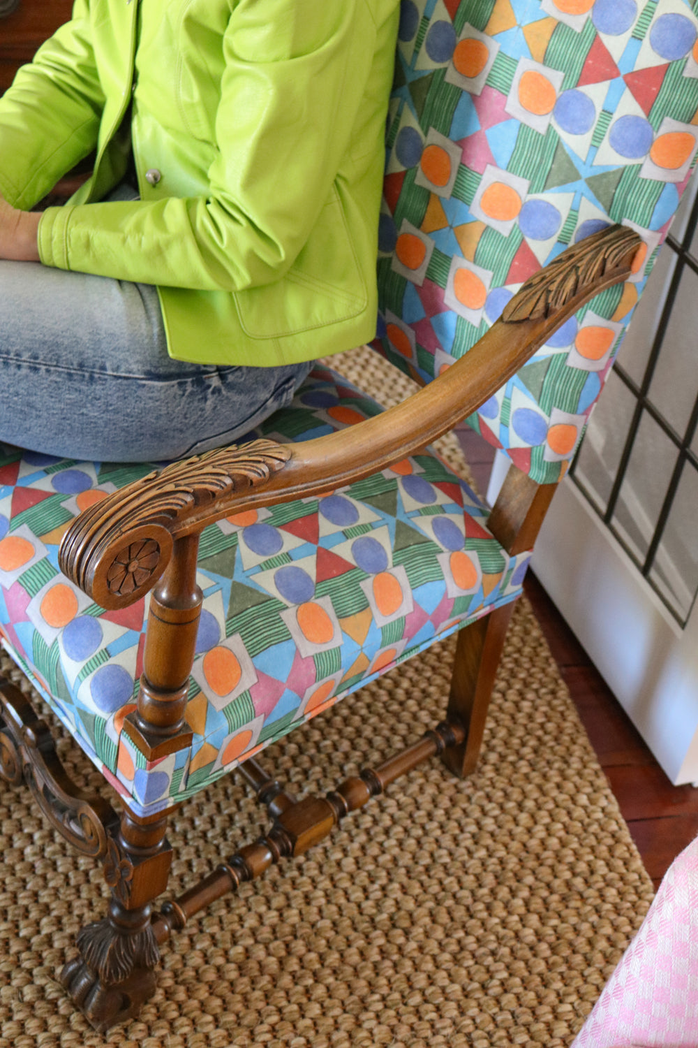 Detail of a seated woman in lime green cardigan and jeans on an ornate wooden side chair with geometric patterned upholstery.